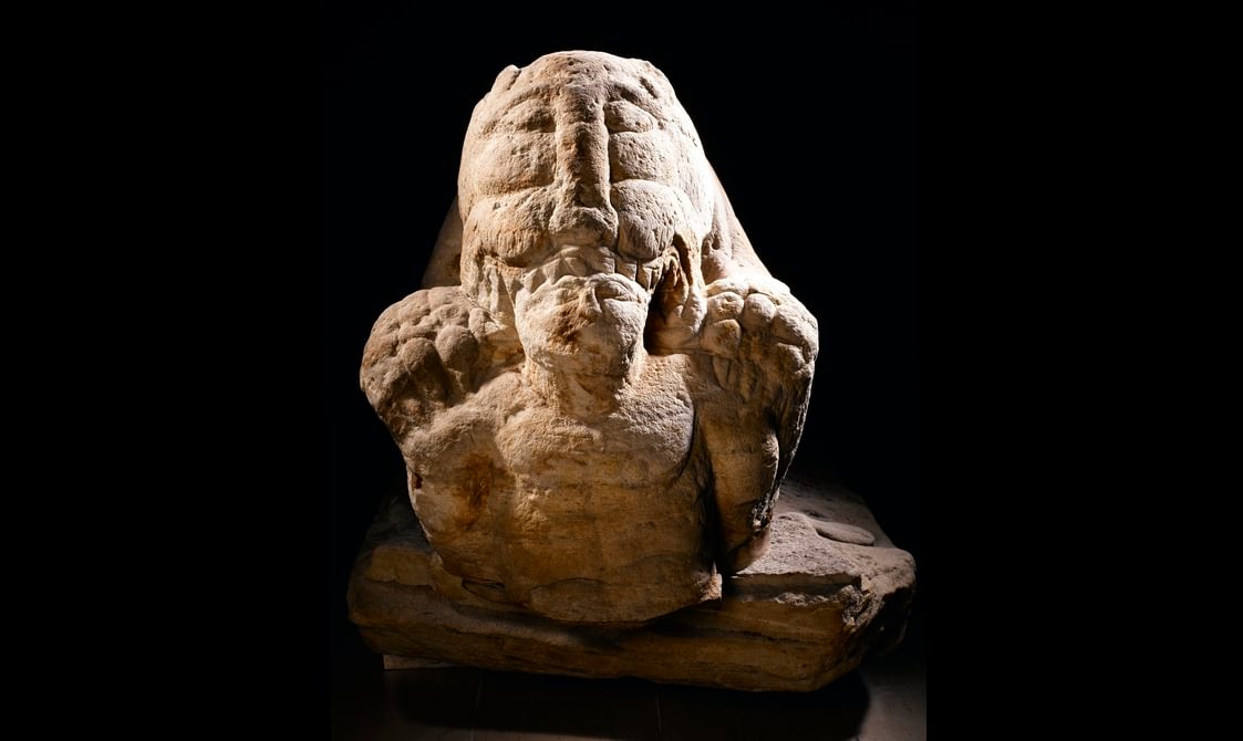 Frontal view of a large, intimidating stone sculpture. Focus is on a man's head and torso being held and devoured by an exaggeratedly monstrous lion.
