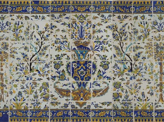 A long, rectangular panel made of hundreds of intricately decorated, painted tiles. This image gives the overall impression of the illustration, which from a distance is of a garden scene with trees all around, and a fountain as the centrepiece. The colours painted are yellow, blue, green and brown.