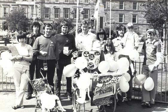 A black and white photo of a group of people standing in a city square holding balloons. There is a child's pram covered in protest signs and buttons.