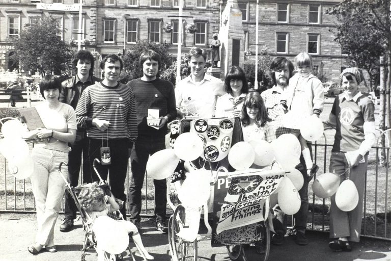 A black and white photo of a group of people standing in a city square holding balloons. There is a child's pram covered in protest signs and buttons.