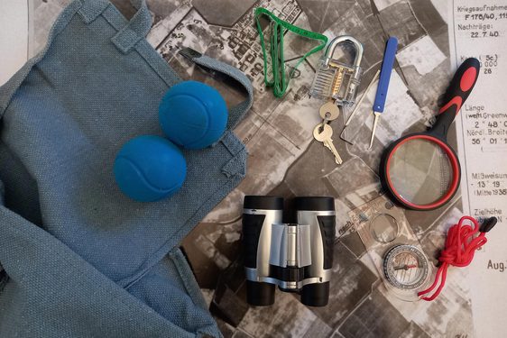 Blue tennis balls, binoculars, magnifying glass, compass and lock and key lie beside blue satchel bag.