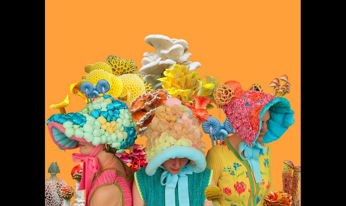 Three perfomers stand in a semi-circle wearing colourful headdresses that resemble mushrooms.