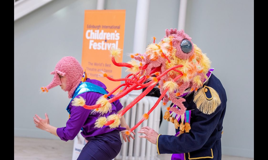 Two performers in a museum gallery. They are wearing colourful, fluffy head costumes that resemble shrimps.