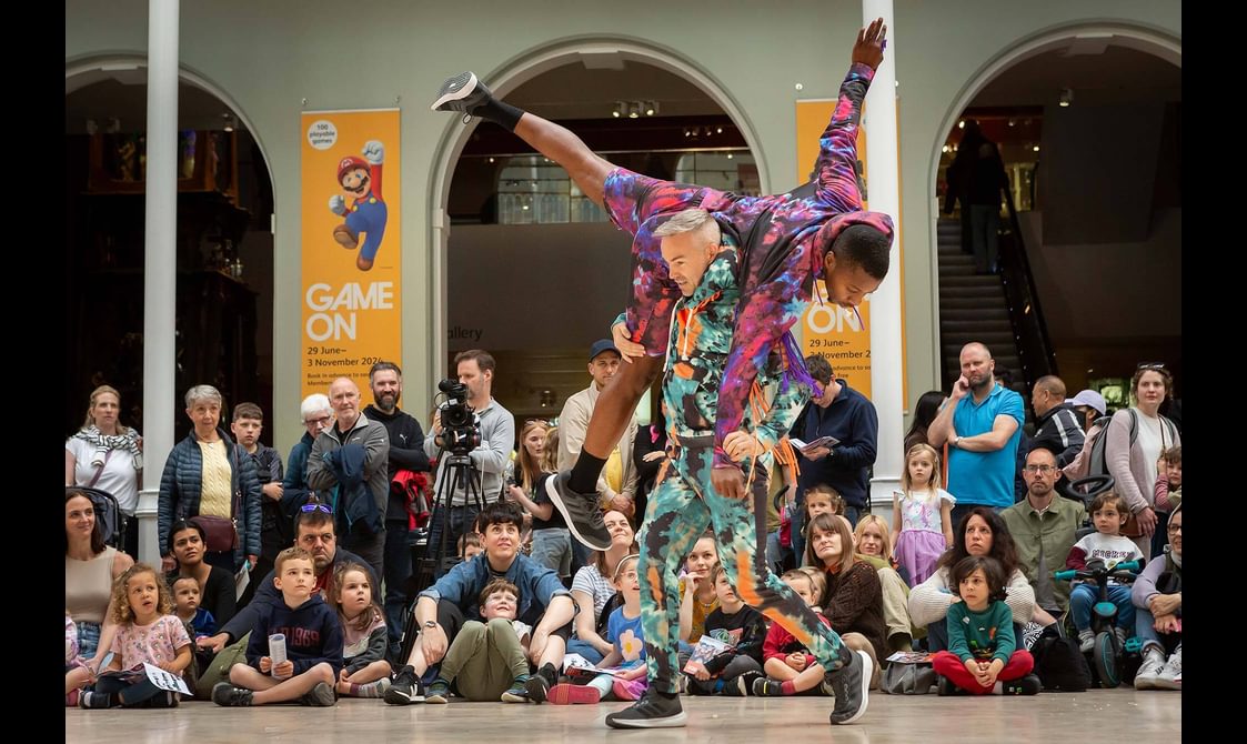 Two dancers perform a lift in front of a large group of people in a museum gallery.