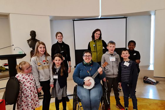 A group of children stand around a person sitting in a wheelchair playing a banjo