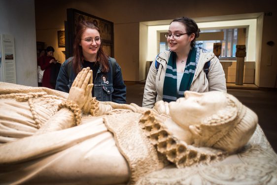 Two visitors in a gallery stand over a statue of a figure lying down with its hands folded in prayer.