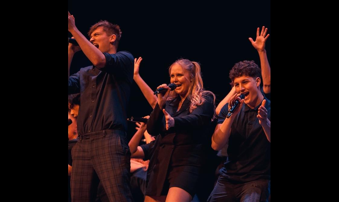 A group of singers wearing black stand on a stage singing into handheld microphones.