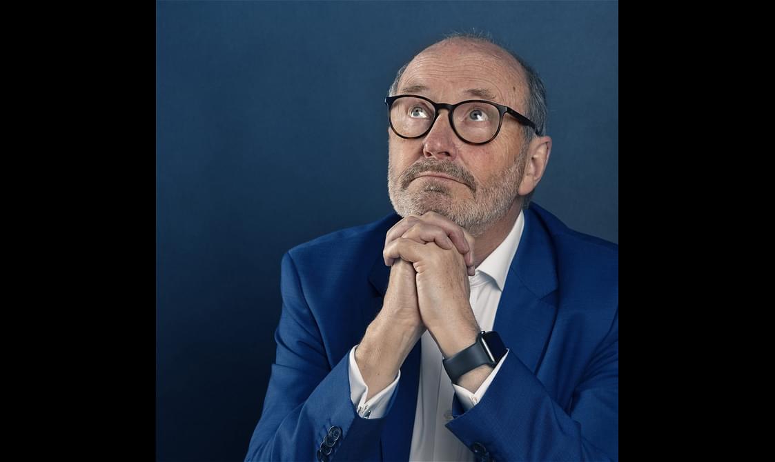 Fred MacAulay wearing black glasses and a blue suit, holding hands together as if in prayer.