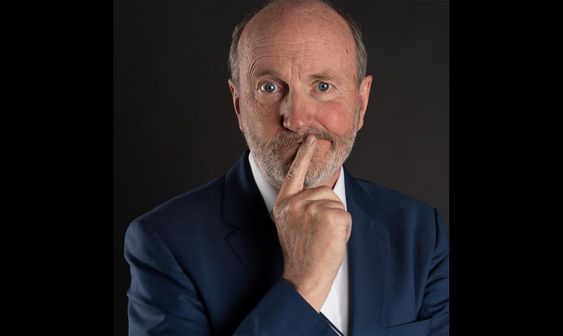 Fred MacAulay in a dark blue suit. His finger is on his chin as if he is questioning something.