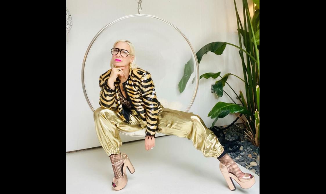 Lu in Luland wearing golded trousers, a gold leopard print jacket, and black glasses. They are sitting in a hanging chair and posing for the camera.