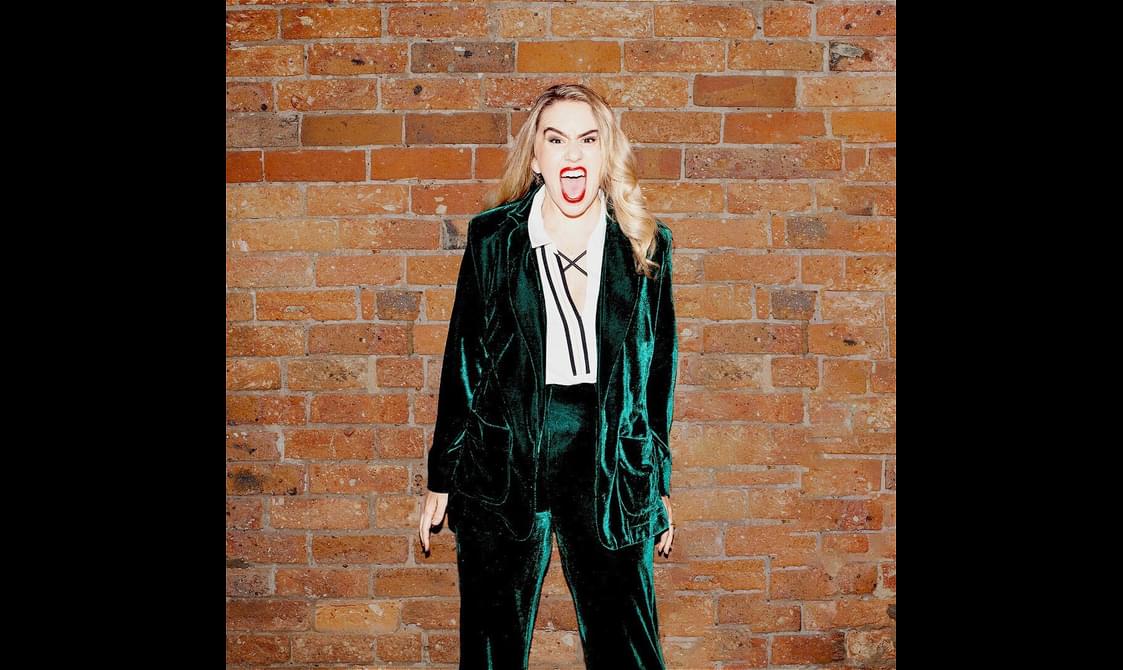 Michelle Brasier wearing a dark green velvet suit standing in front of a brick wall.