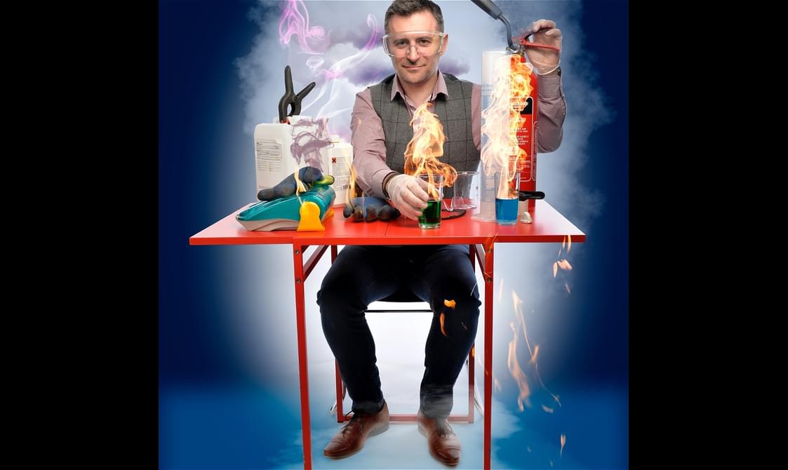 Mark Thompson sits in jeans and a waistcoat at a red table. Flames and smoke spread across the table as a result of science experiments.
