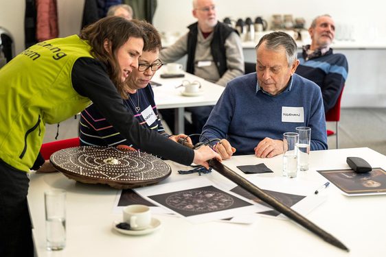 Two people sitting at a table listening to a member of museum staff talk about a sword from the collections.