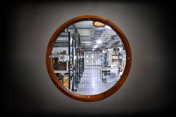 A small circular window, like those you would see in the hull of a ship, gives a glimpse into a museum storage area filled with racks containing boxes and folders.