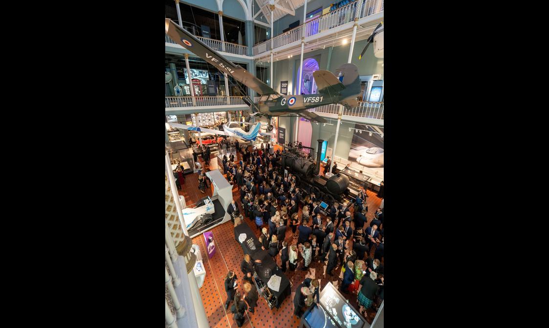 A large group of people at a drinks reception in a multi-level museum gallery. There are different vehicles on display in the gallery and aircrafts suspended from the ceiling.