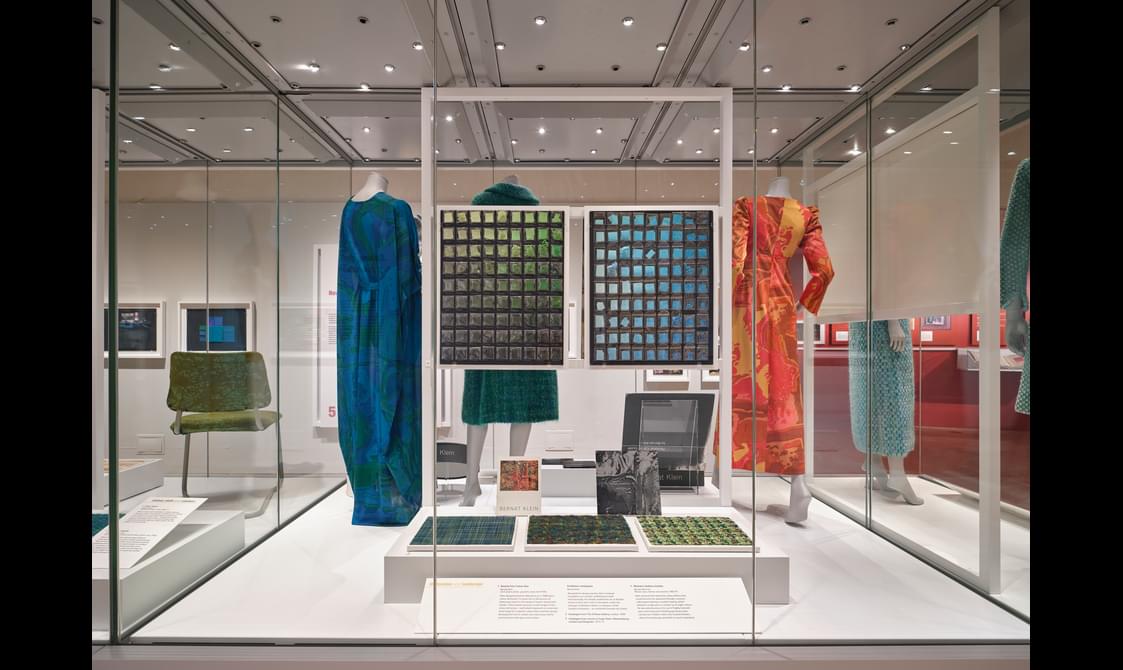A glass exhibition case with an information board, material samples and 3 clothed mannequins.