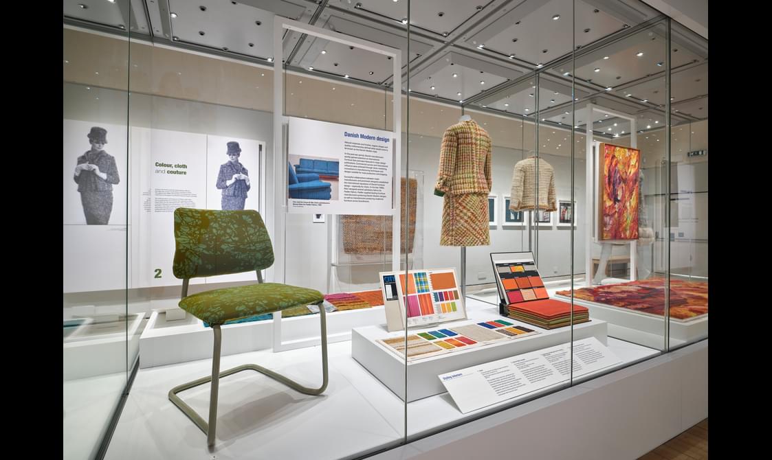 A glass exhibition case with a green chair, information boards, material samples and 2 clothed mannequins.
