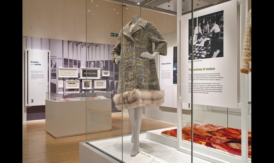 A glass exhibition case with an information board, rug and clothed mannequin.