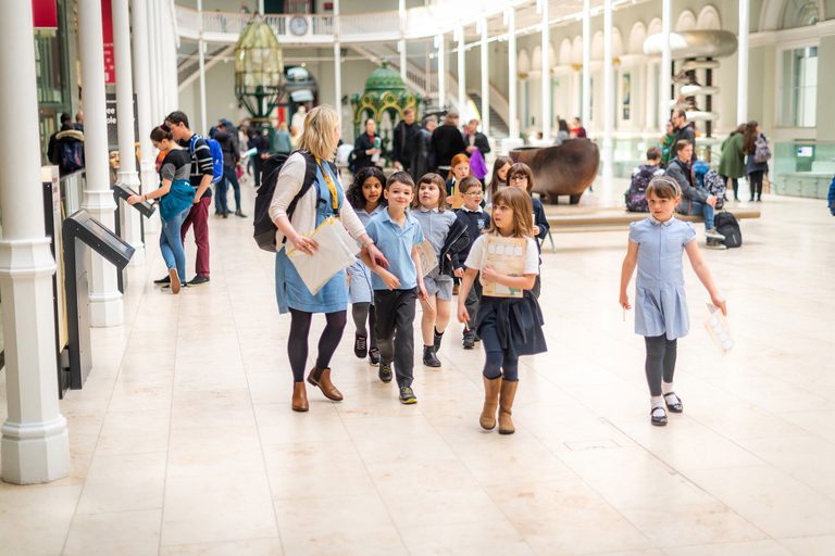 A teacher leads a group of young school children through a large, bright, open gallery.